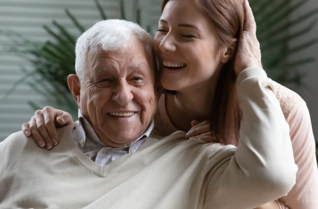 An older adult smiles while their adult child hugs them from behind during a visit in senior living