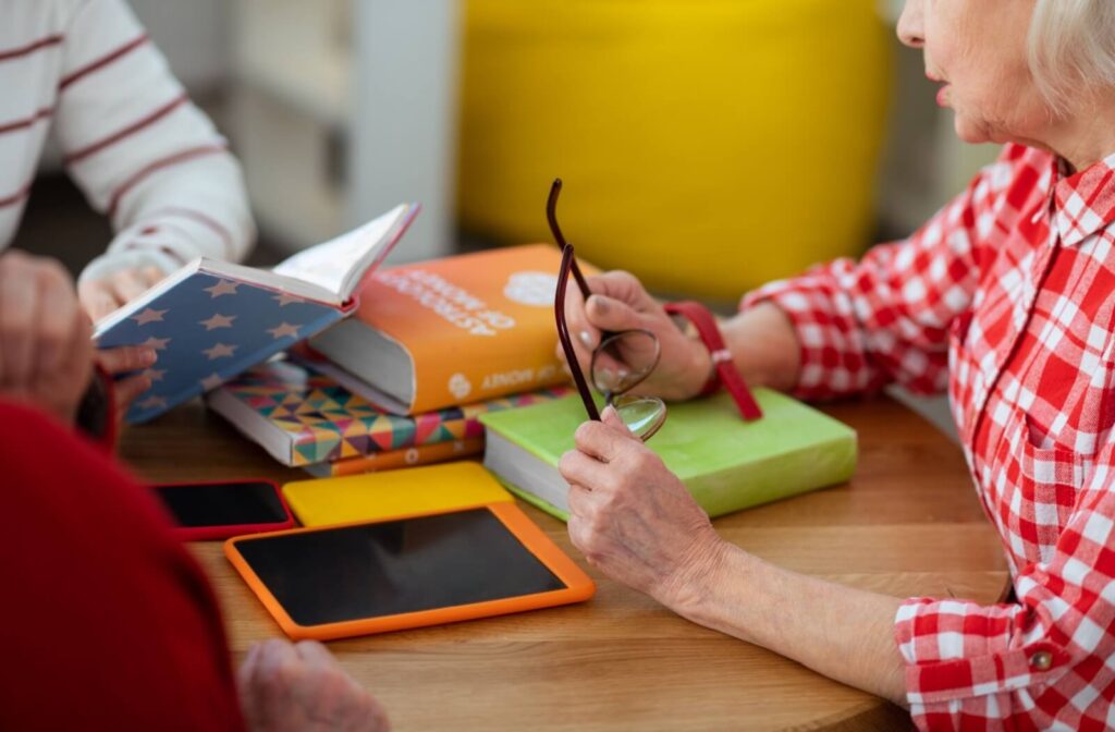A photo of 3 seniors sitting around a table, with books and tablets in the middle. One senior is holding a pair of glasses and speaking